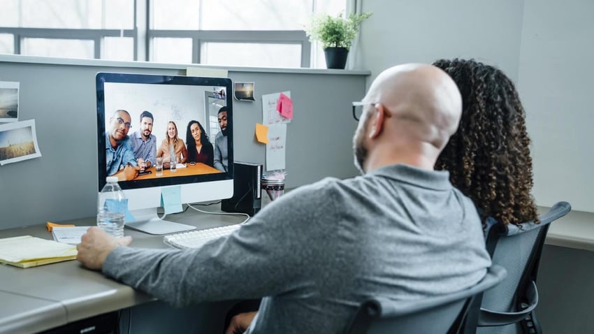 video conference call between two teams a man and a woman in a cubicle looking at a monitor where they are holding a video conference call with another professional team